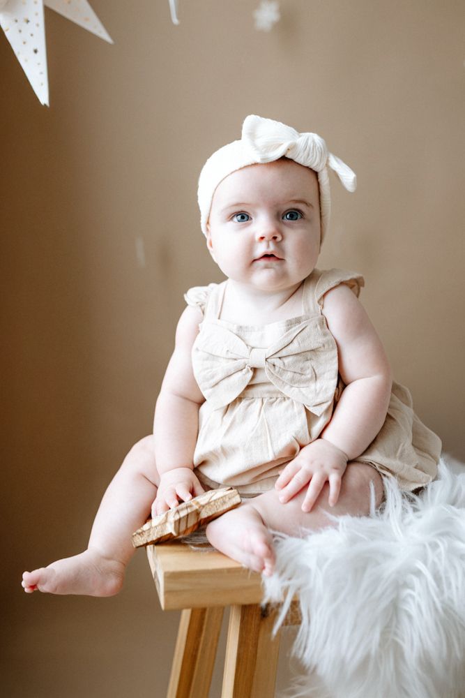 Baby wearing a cream bow headband and dress sits on wooden stool with white fur under hanging paper stars.