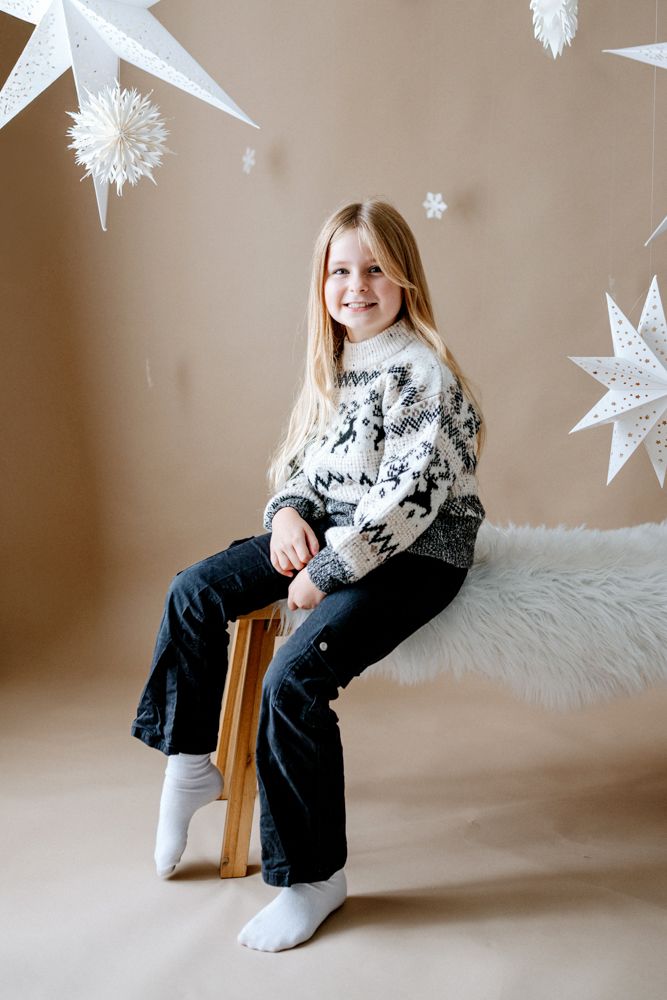 A child poses in a cozy Nordic sweater on a wooden chair with white star decorations in the background.