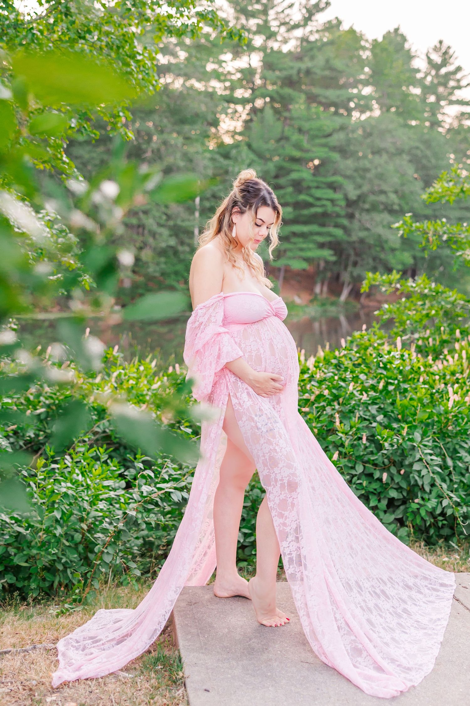 A flowing pink lace maternity gown with off-shoulder design is captured in an ethereal outdoor garden photoshoot.