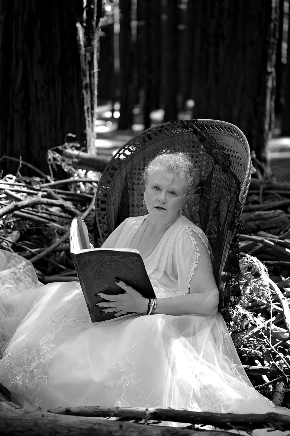 A vintage style black and white photo of someone resting in a white dress on leaves and branches in a forest setting.
