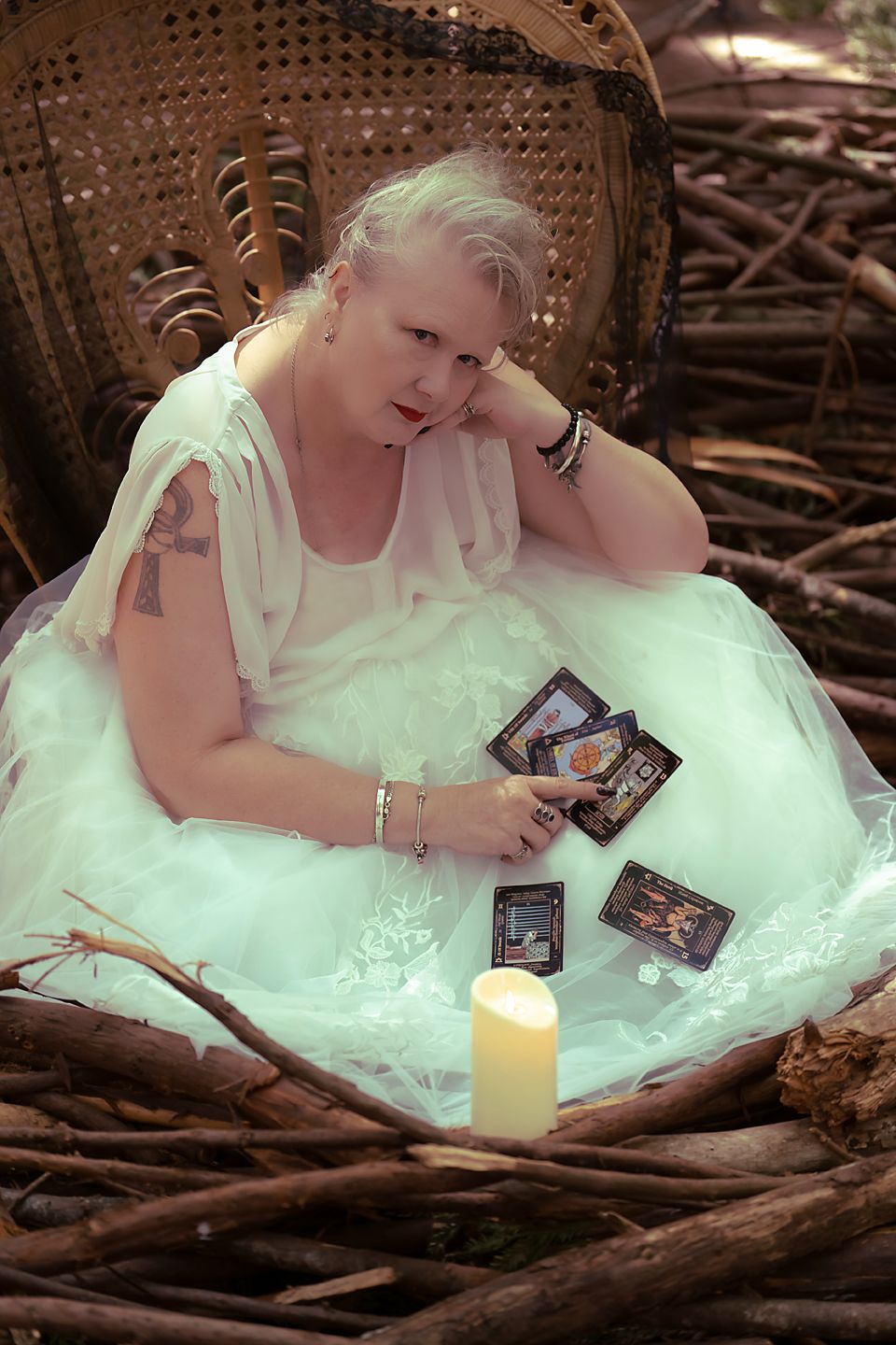 Person in a white dress sits on forest ground reading tarot cards by candlelight in an enchanted woodland setting.