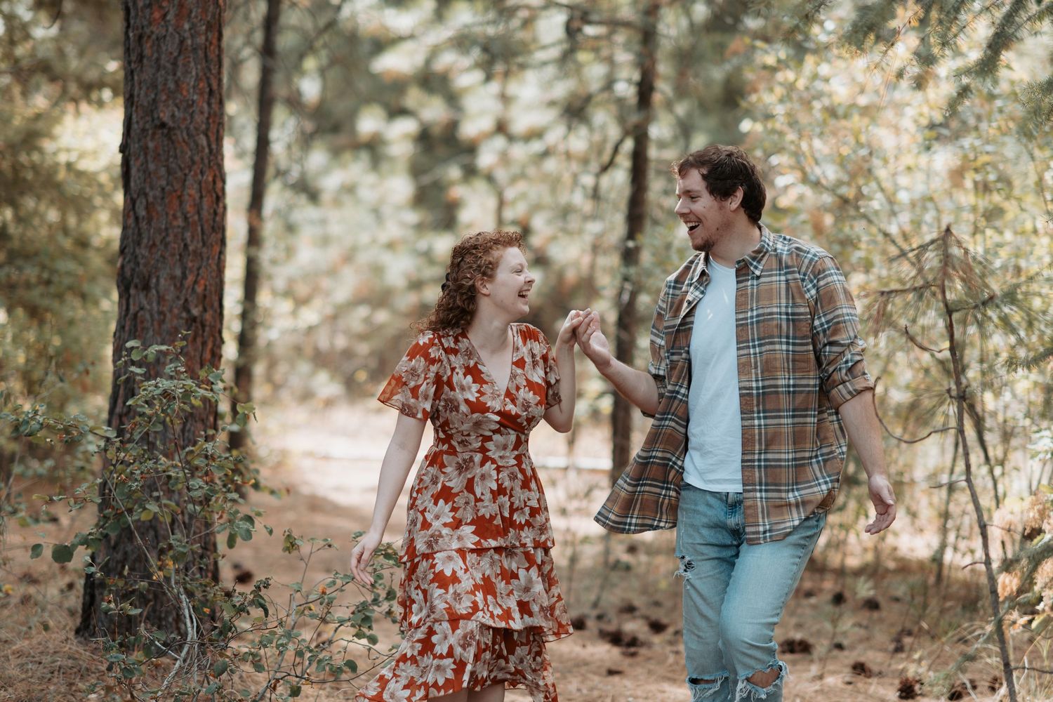 Young couple holding hands winding through the pine trees in Post Falls Idaho during their playful engagement session.