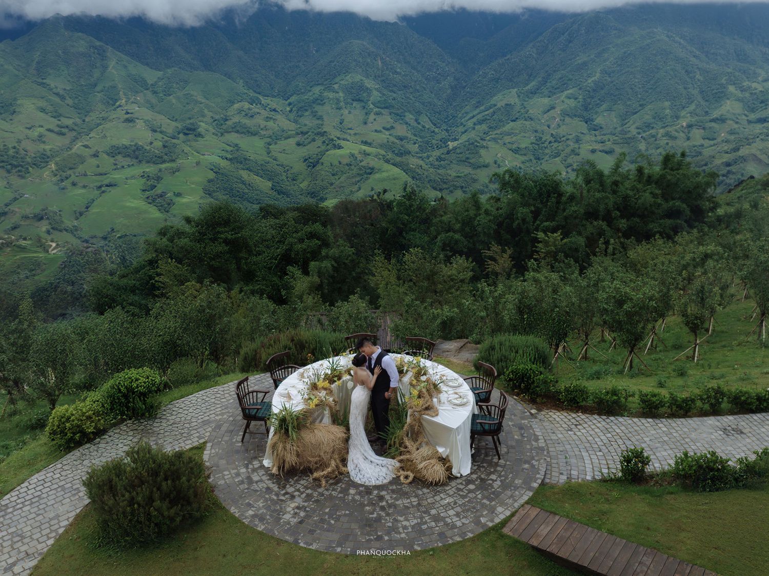 Aerial view of bride and groom at a decorated outdoor wedding table surrounded by lush green mountain landscape.