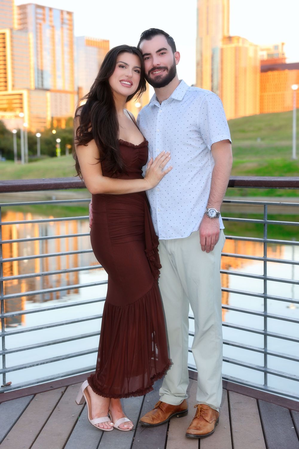 A couple poses together on a deck at sunset with city buildings in the background.