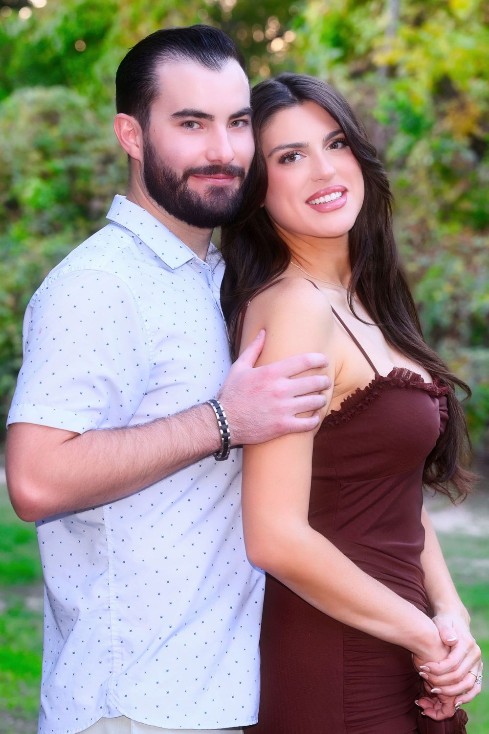 A couple in burgundy dress and white polka dot shirt embrace outdoors against a green natural background.