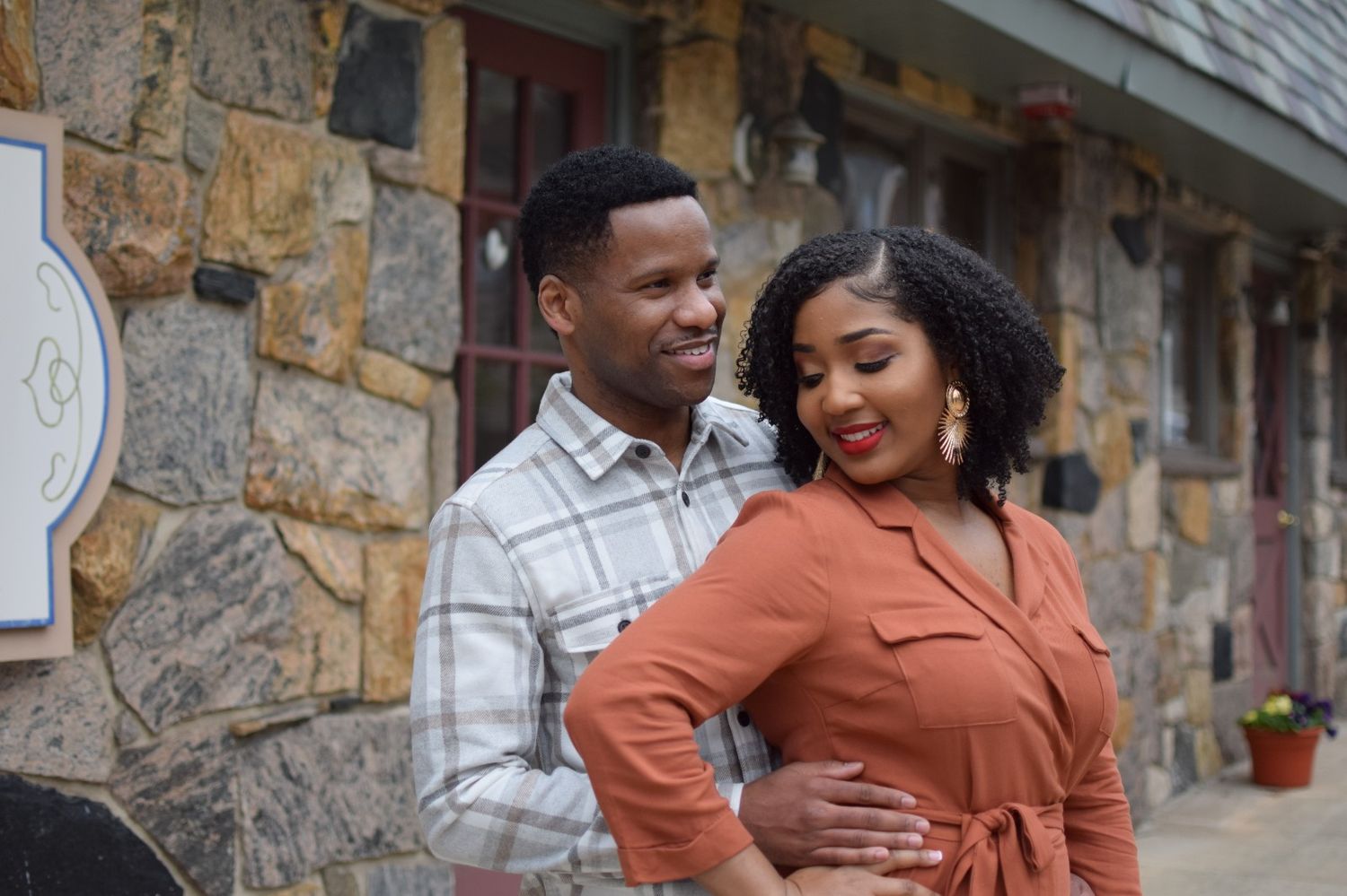 A stylish couple standing close together outdoors, with him wearing a plaid shirt and her in a rust-colored top.
