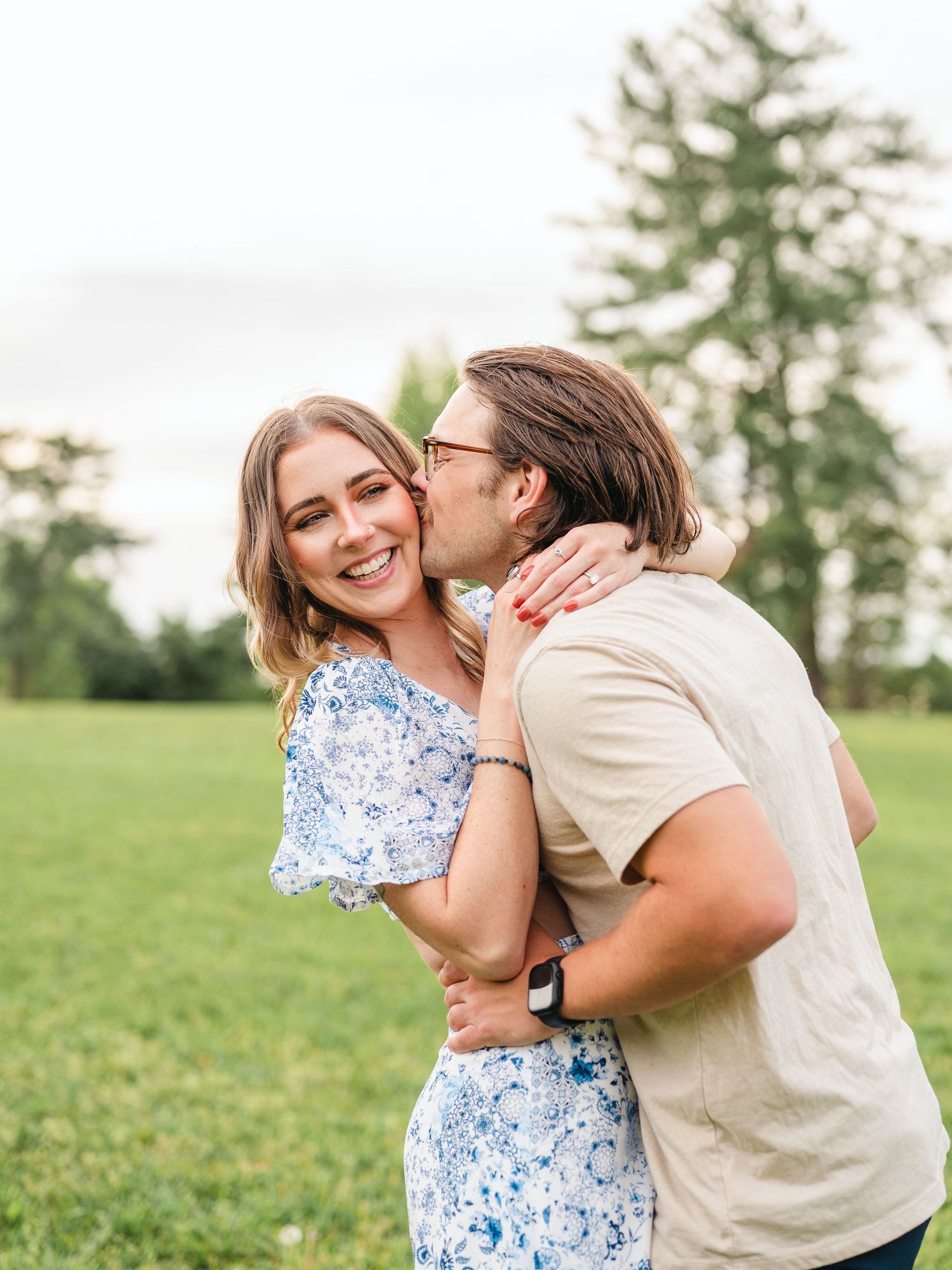 A joyful couple embraces in a grassy field with tall evergreen trees in the background on a sunny day.