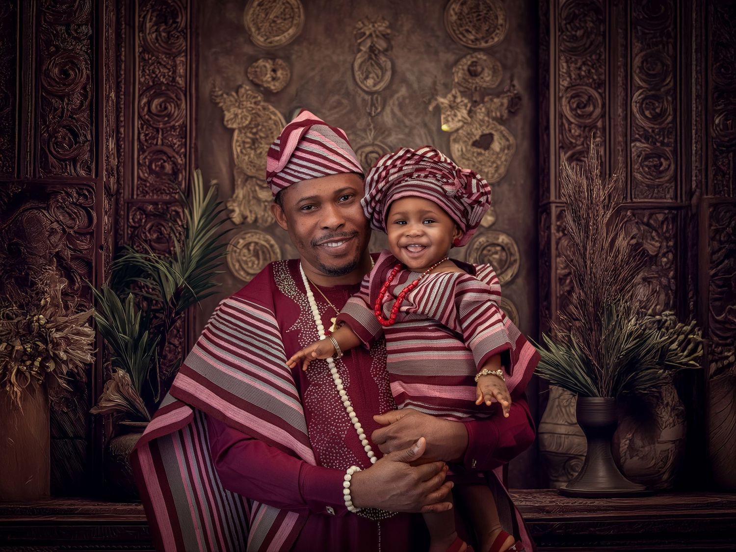Woman in vibrant burgundy traditional dress and headwrap poses joyfully against ornate wooden carved wall panels.