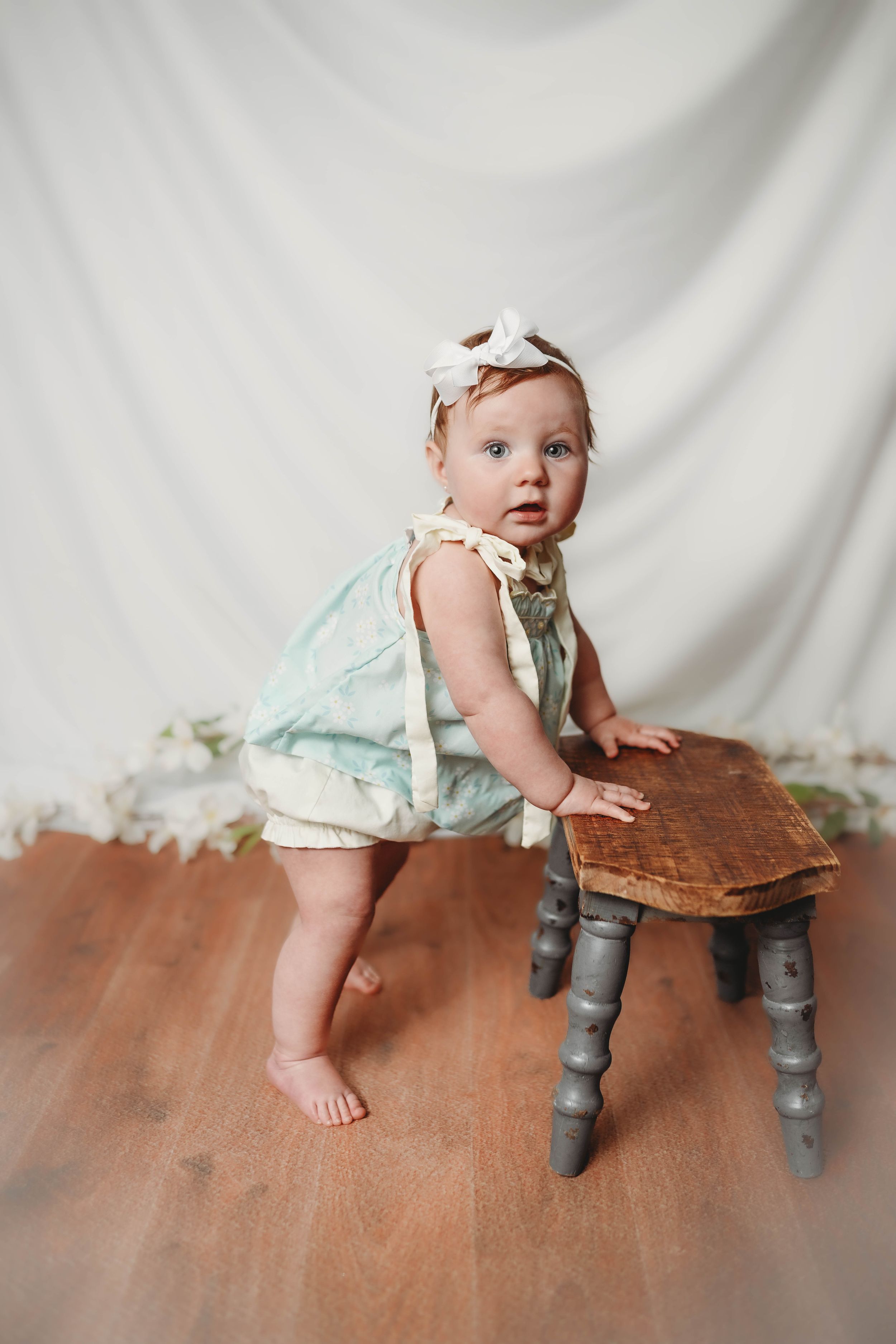 Baby in mint dress and bow headband stands next to vintage wooden stool.