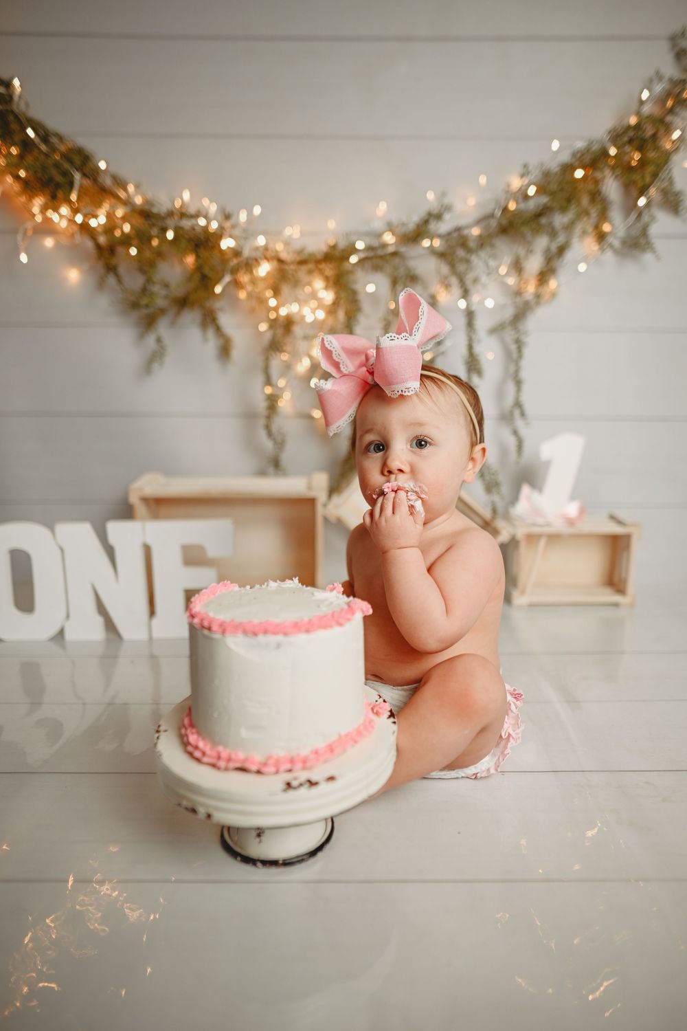 Baby in pink bow sits next to white birthday cake with pink trim under twinkle light garland.