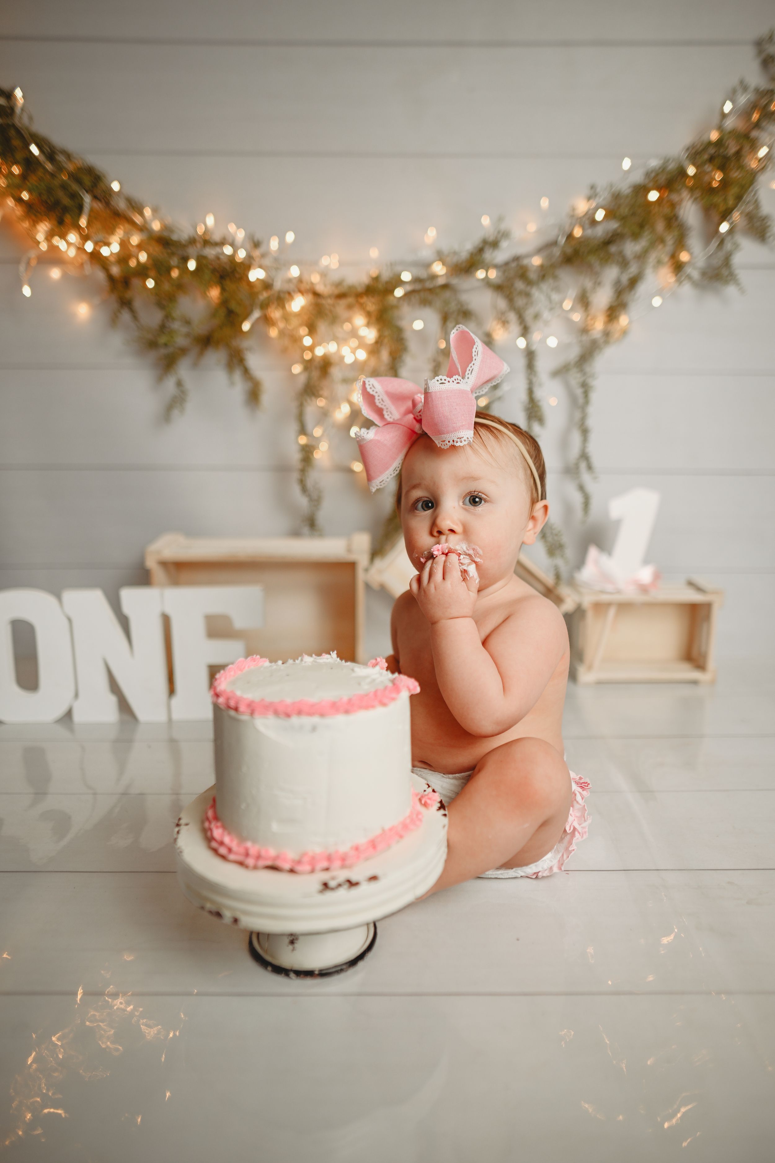 Baby in pink bow sits next to white birthday cake with pink trim under twinkle light garland.
