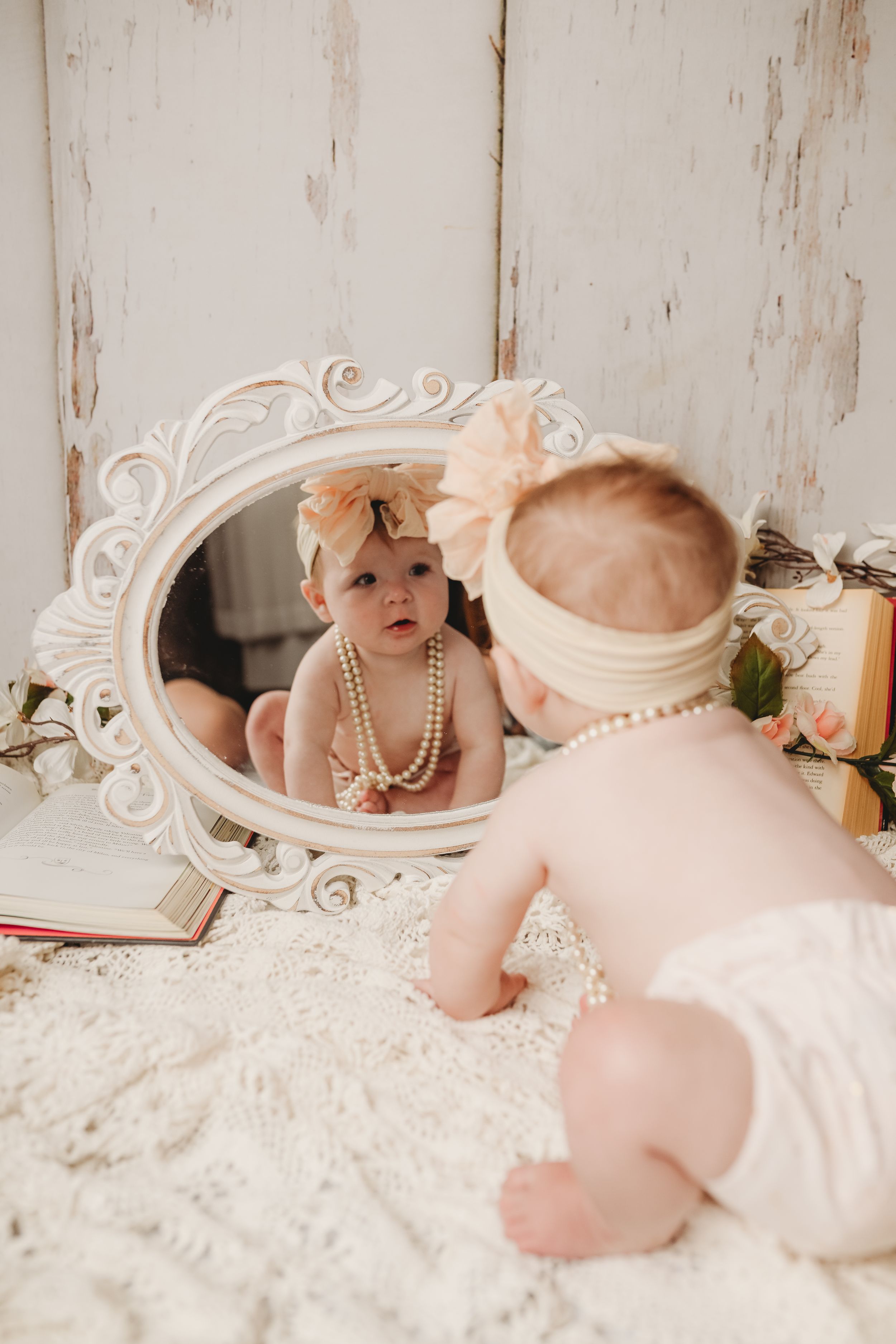 Baby wearing pearls and bow looks at reflection in ornate white mirror on lace blanket.