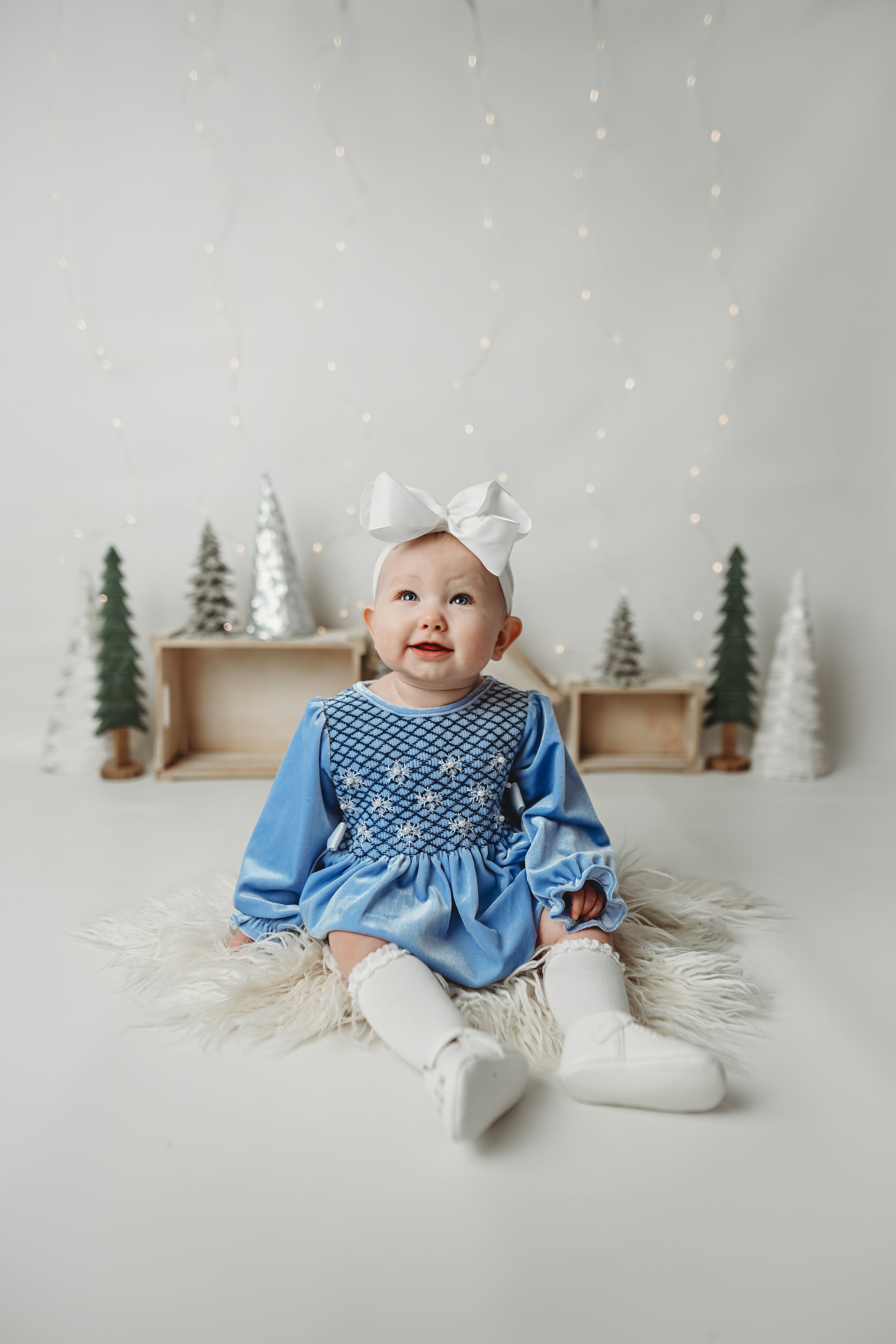 Baby in blue smocked dress sits on fur rug with winter decorations and trees in background.