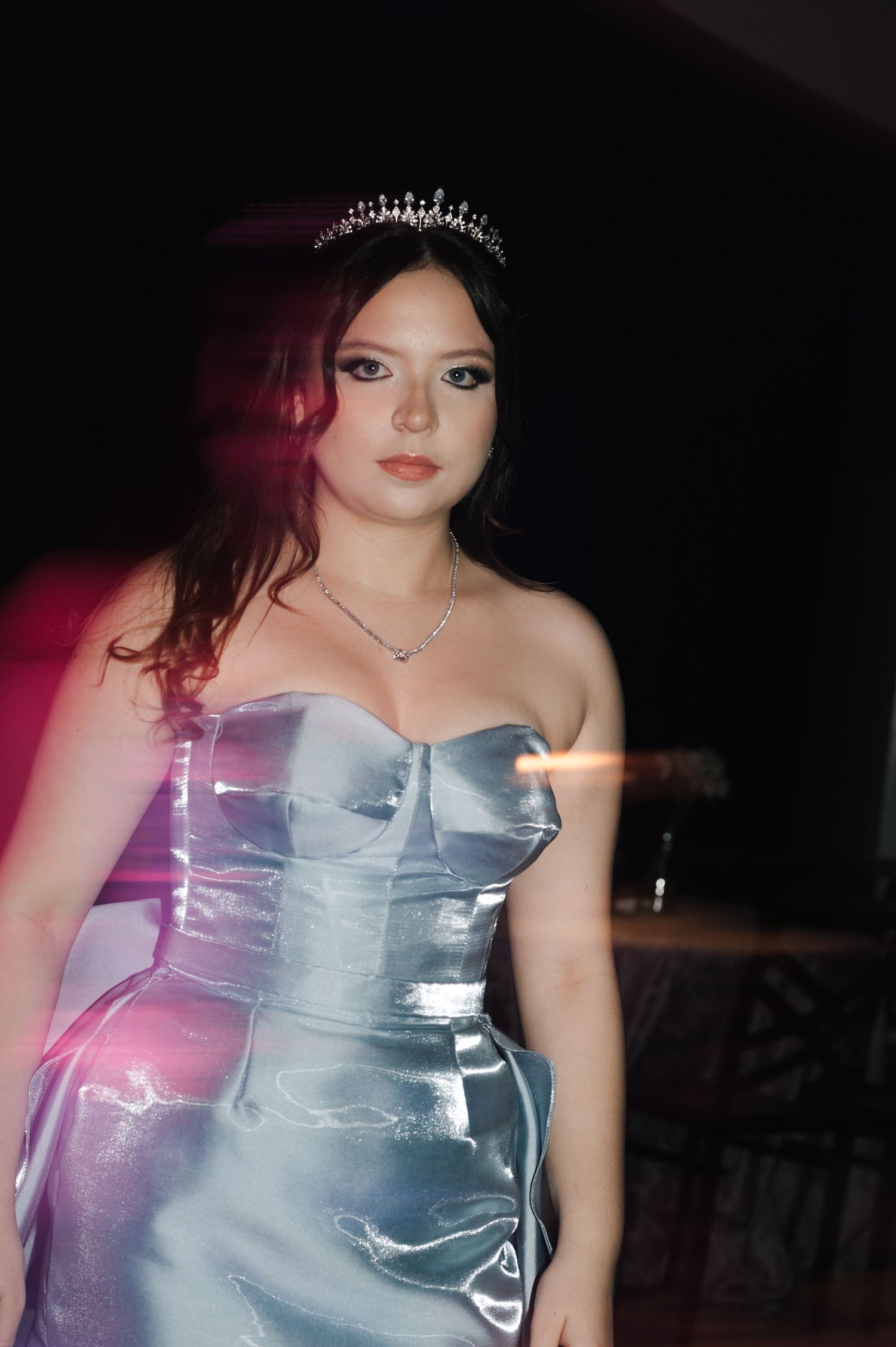 A person in a metallic silver strapless dress poses against a dark background with pink lighting effects.