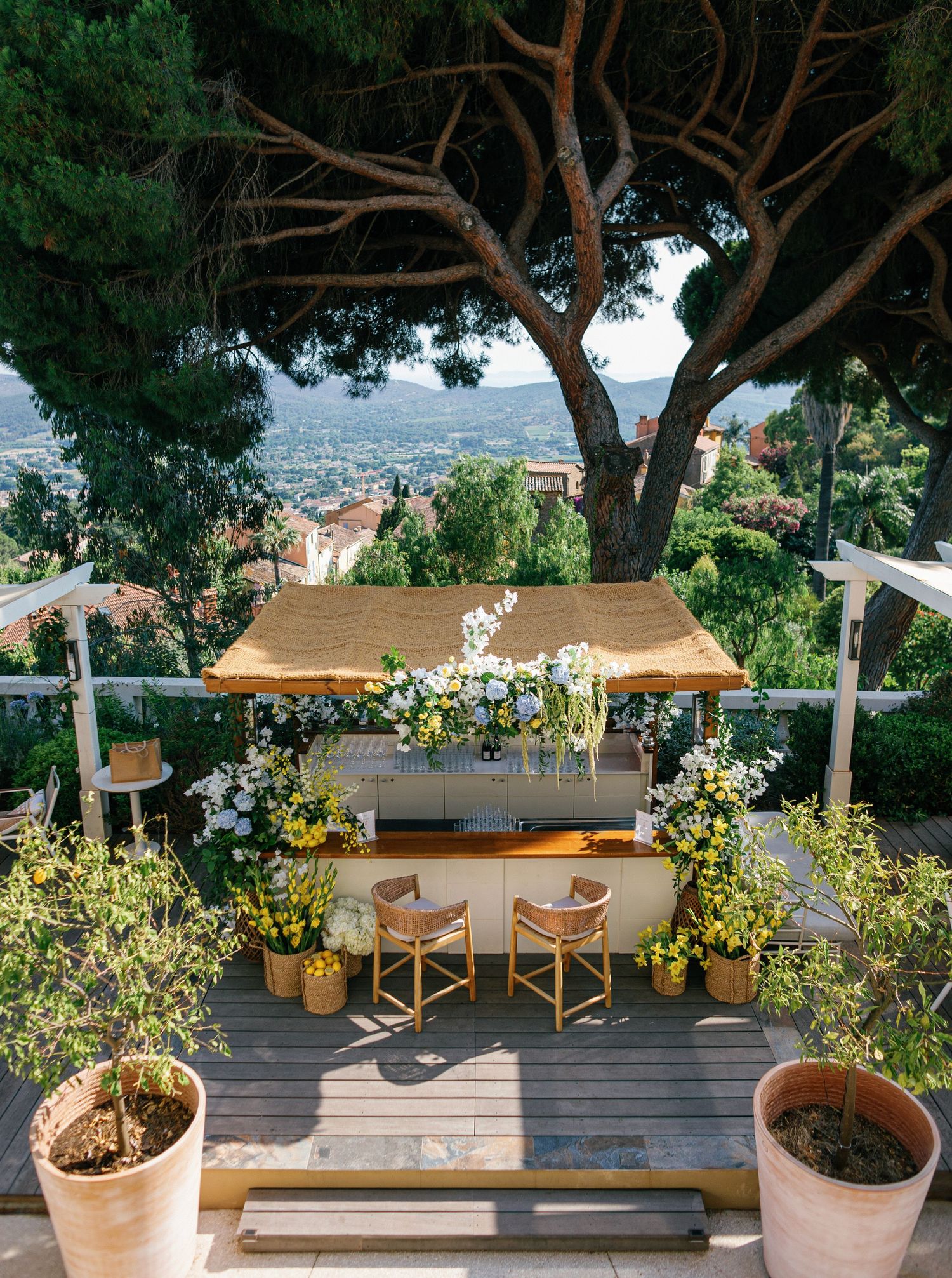 A wooden deck terrace with built-in benches and seating areas surrounded by potted plants overlooking a coastal view.