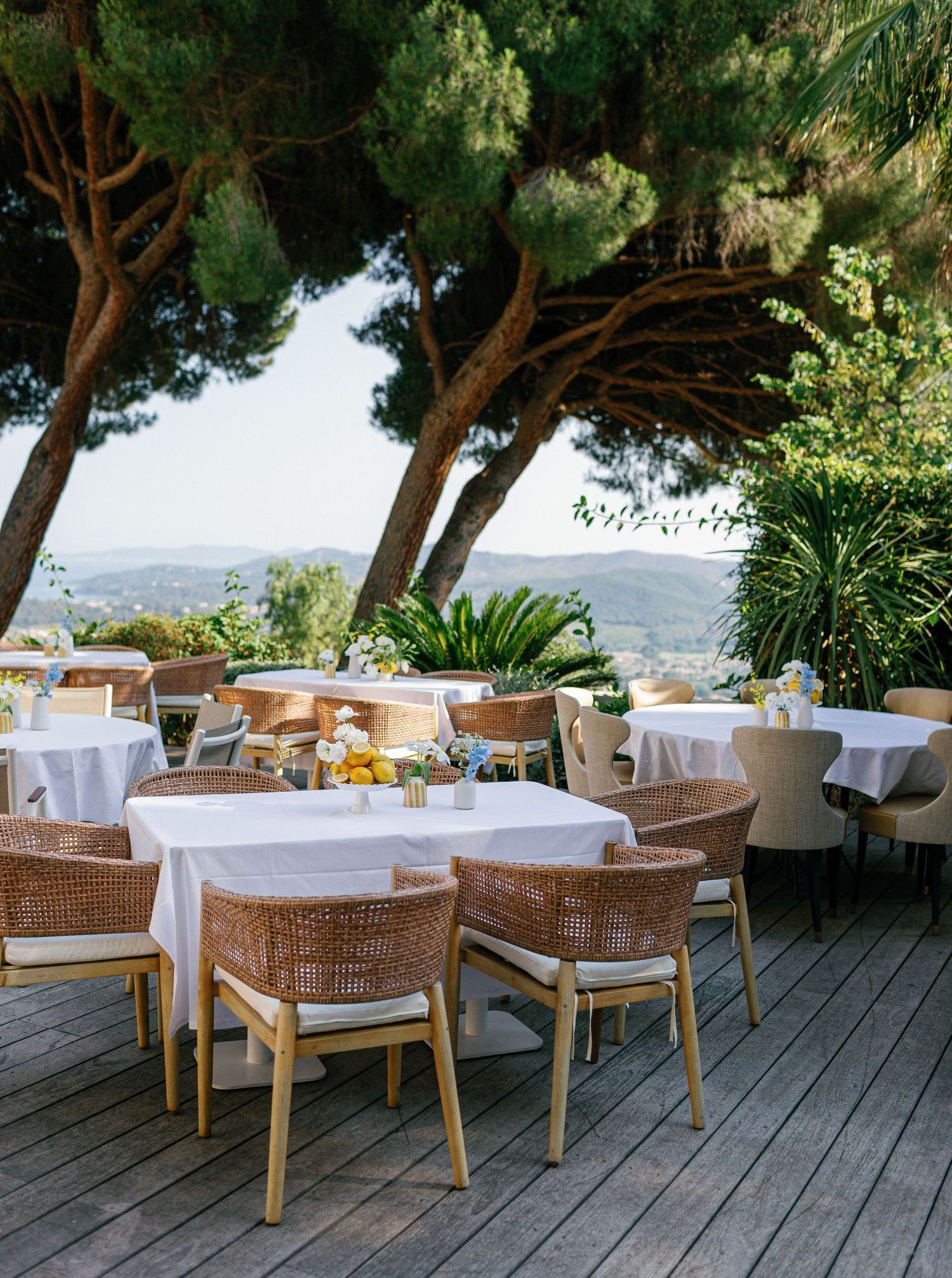 Elegant outdoor restaurant terrace with white tablecloths and wicker chairs overlooking scenic mountain views through pine trees.