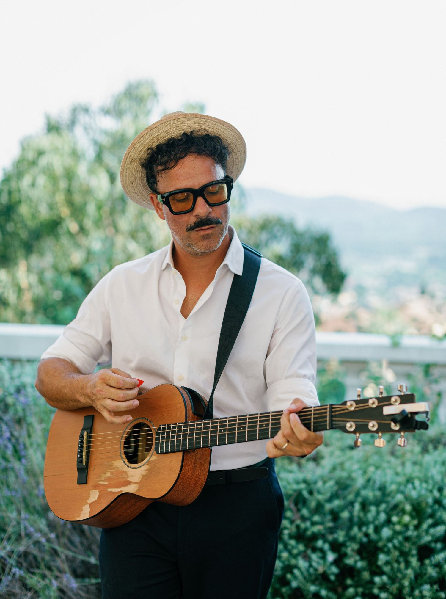 Musician in straw hat and white shirt playing acoustic guitar outdoors with greenery and mountains in background.