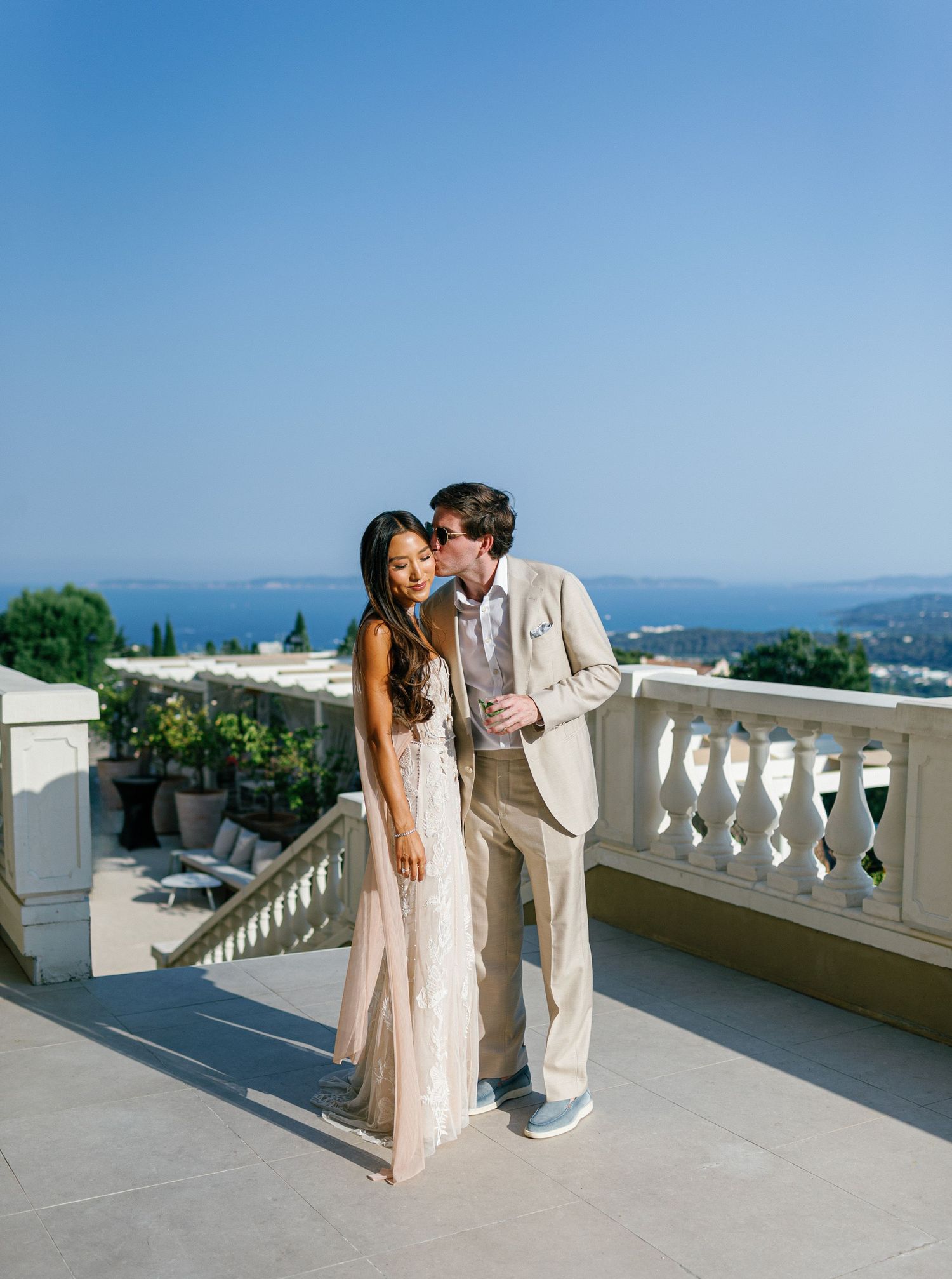 A couple in elegant beige attire share an intimate moment on a Mediterranean balcony with scenic coastal views  at Eden Rose Grand Hôtel.