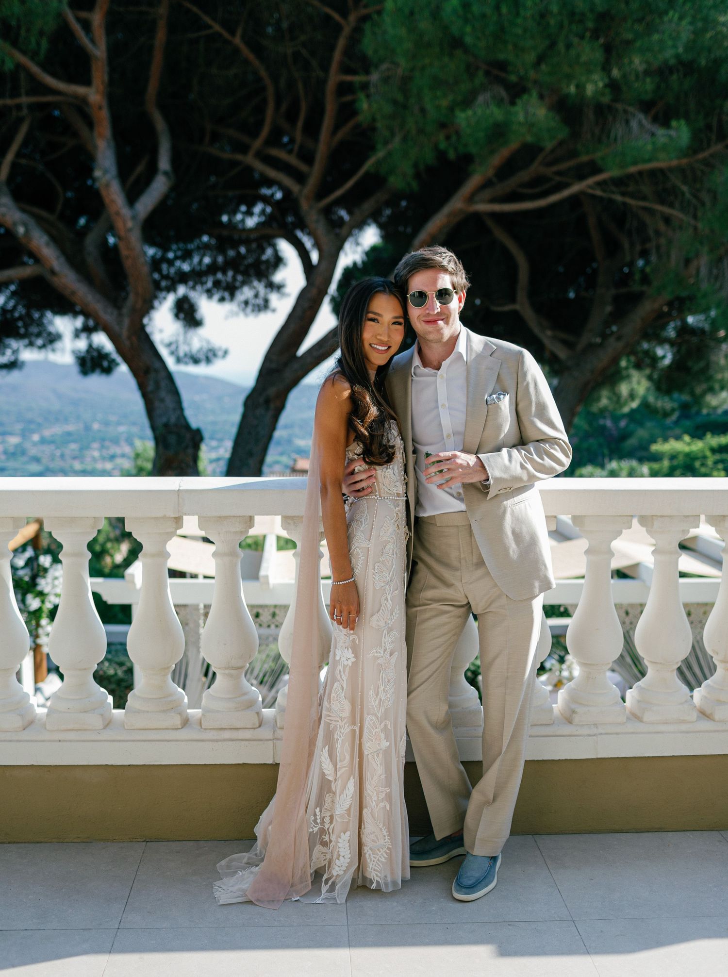 Elegant couple in beige and blush attire pose gracefully against a white balustrade overlooking a scenic landscape.