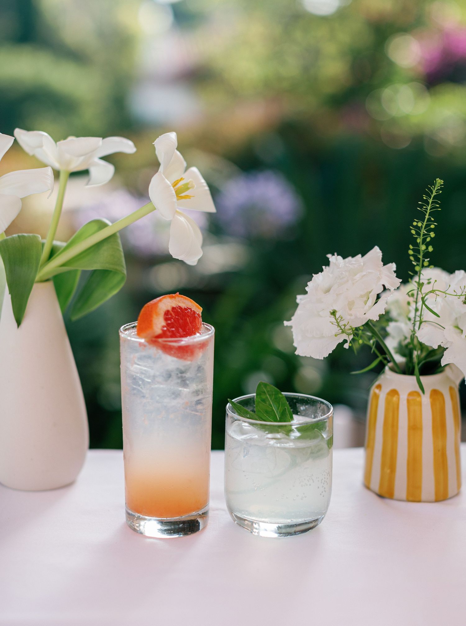 A collection of elegant cocktails with floral garnishes and crushed ice lined up on a white surface in soft natural light.