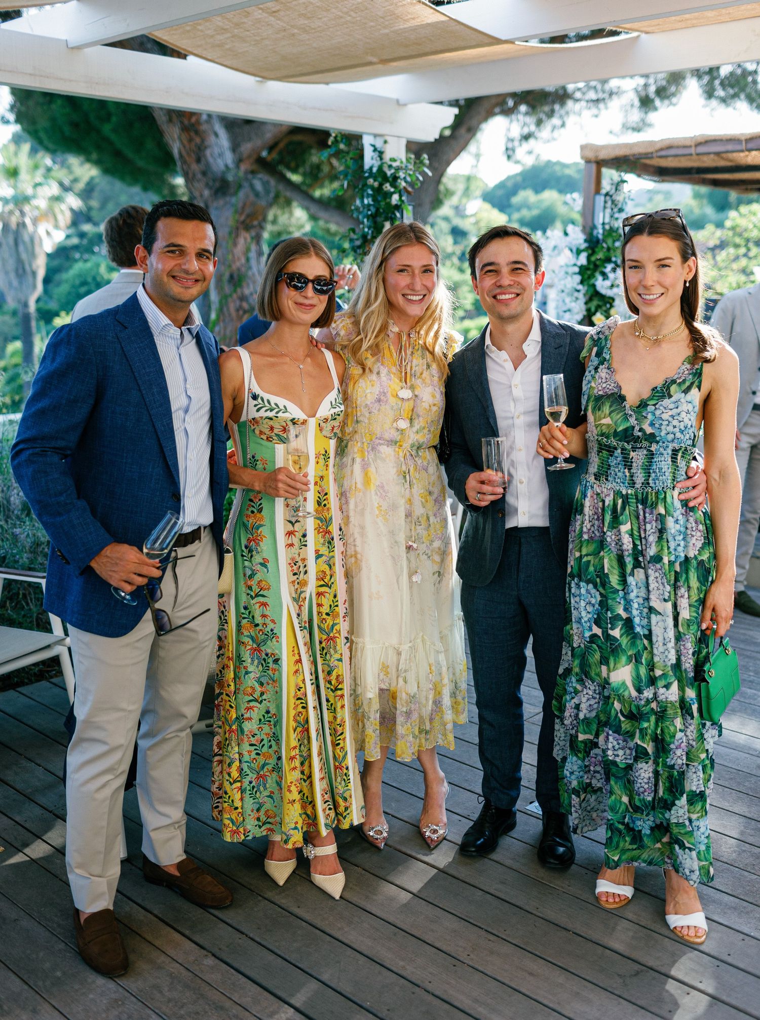 Group of wedding guests in blue suits and floral dresses posing together at an outdoor venue during daytime celebration.