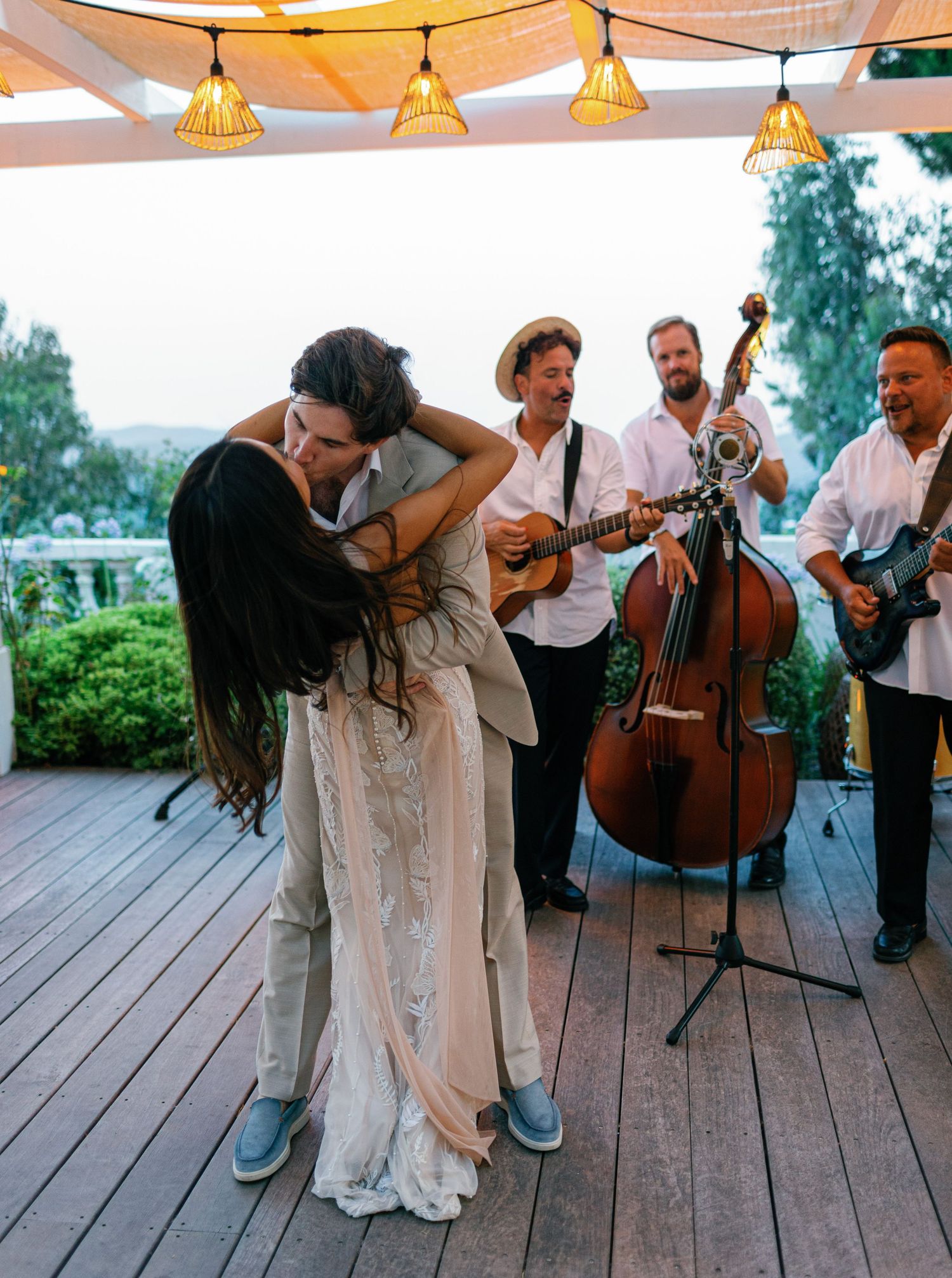 Musicians with guitars and cello perform at an outdoor wedding celebration while a couple dances on a wooden deck.