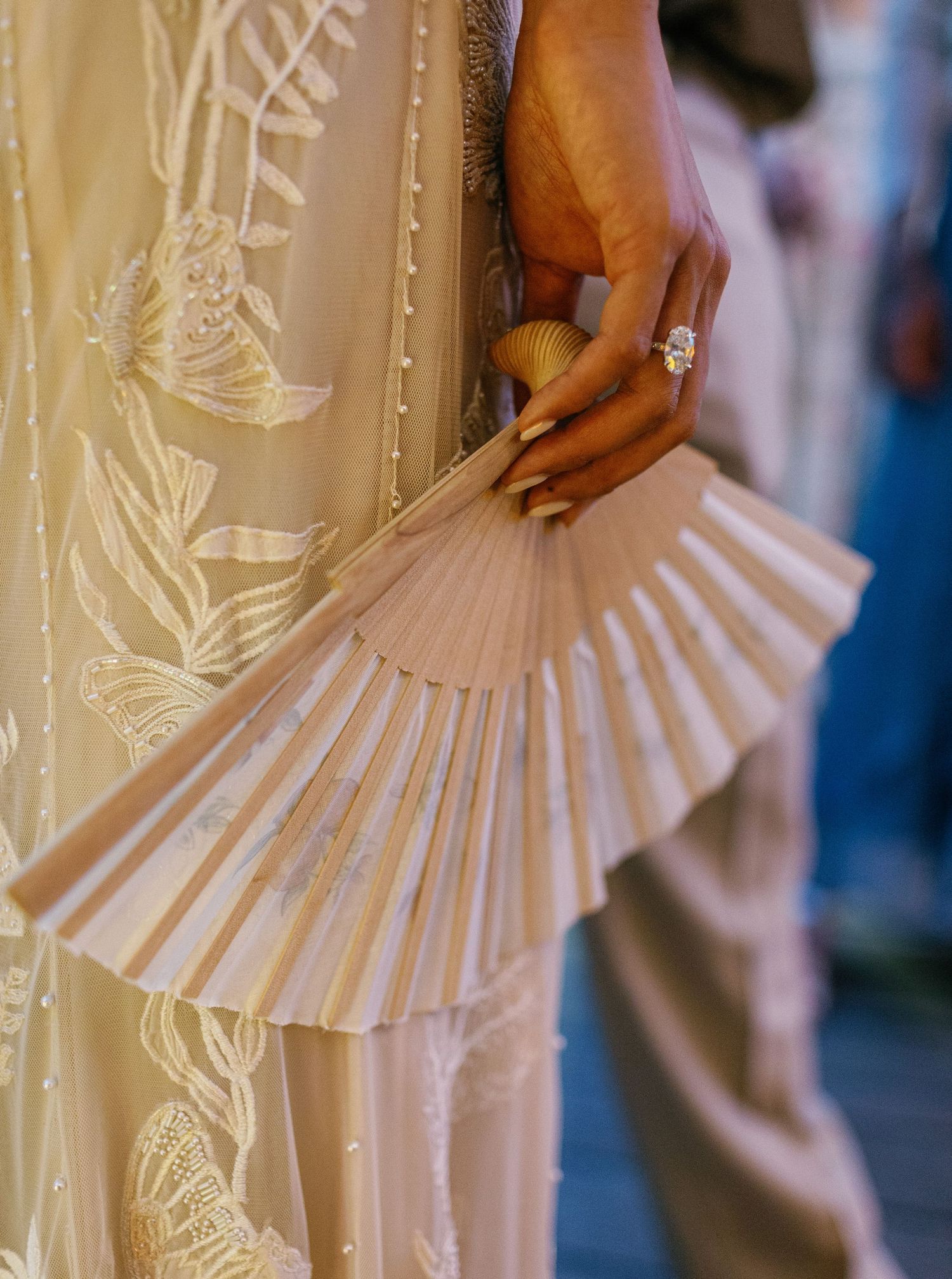 A hand holding an elegant white pleated paper fan against a floral embroidered dress with ornate details.