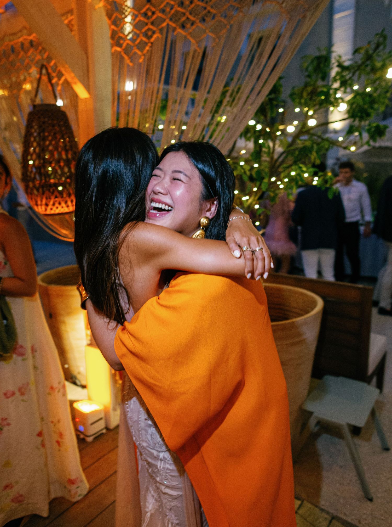 A person in a vibrant orange sweater shares a joyful hug and laugh with another person in a warmly lit indoor space.