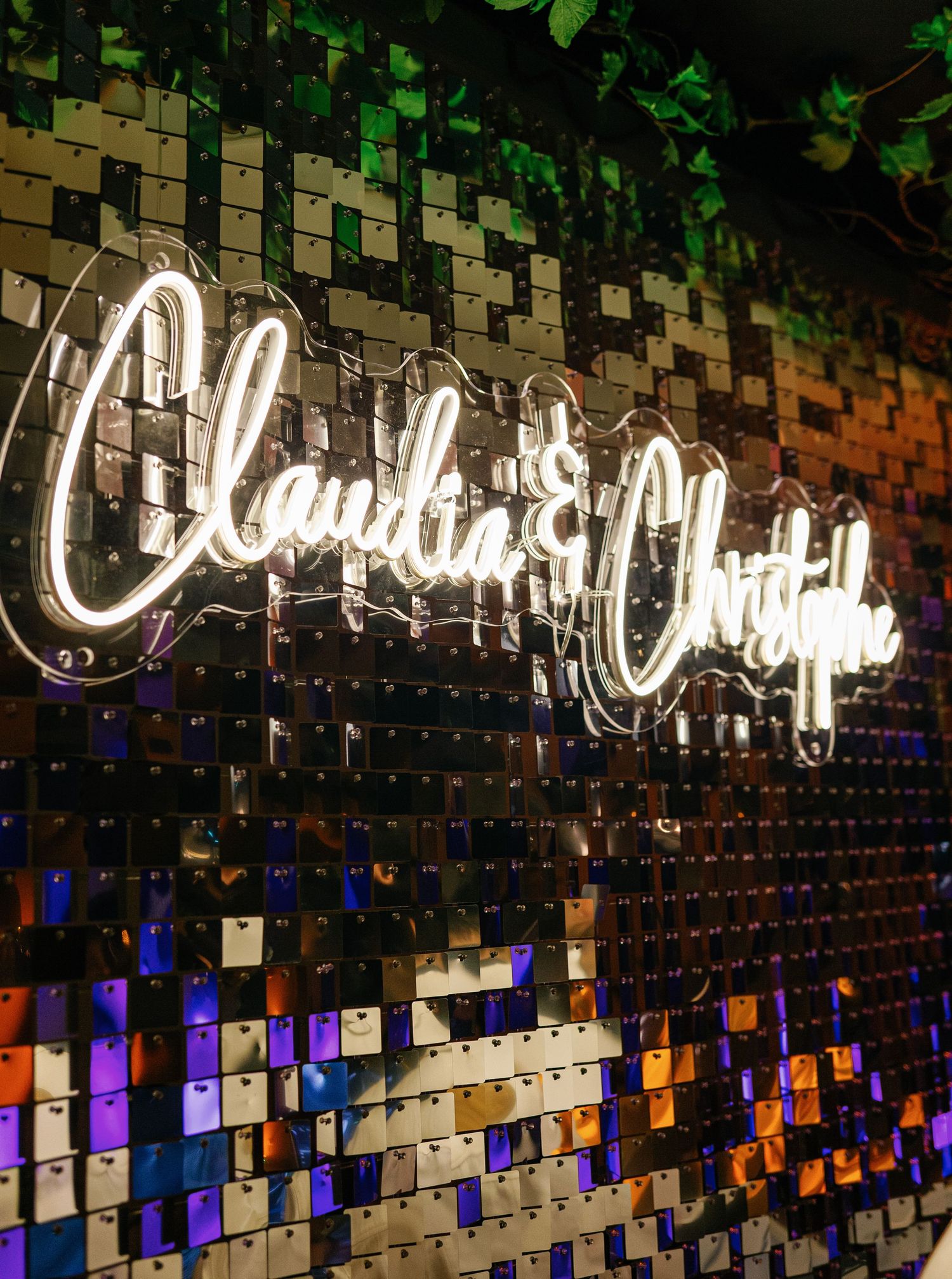 Illuminated neon cursive text glows against a dark mosaic wall with colorful tiles arranged in a geometric pattern.