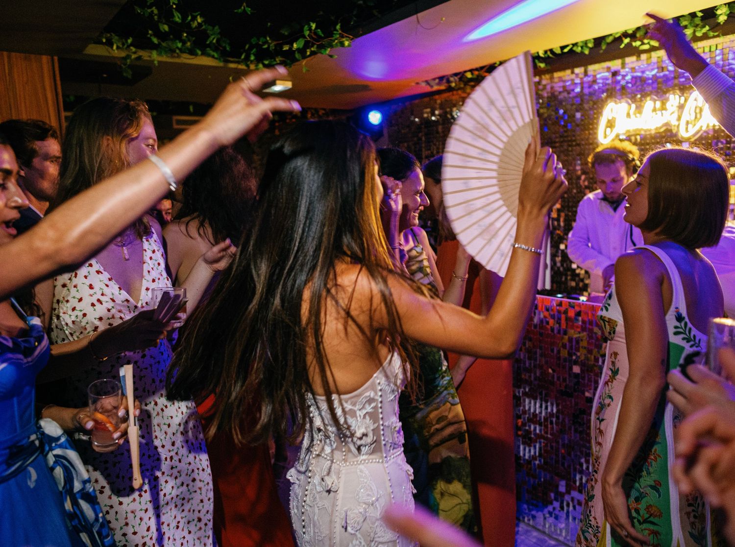 Guests and the bride are dancing and having fun with decorative fans at a vibrant party with purple lighting and sparkly decor.