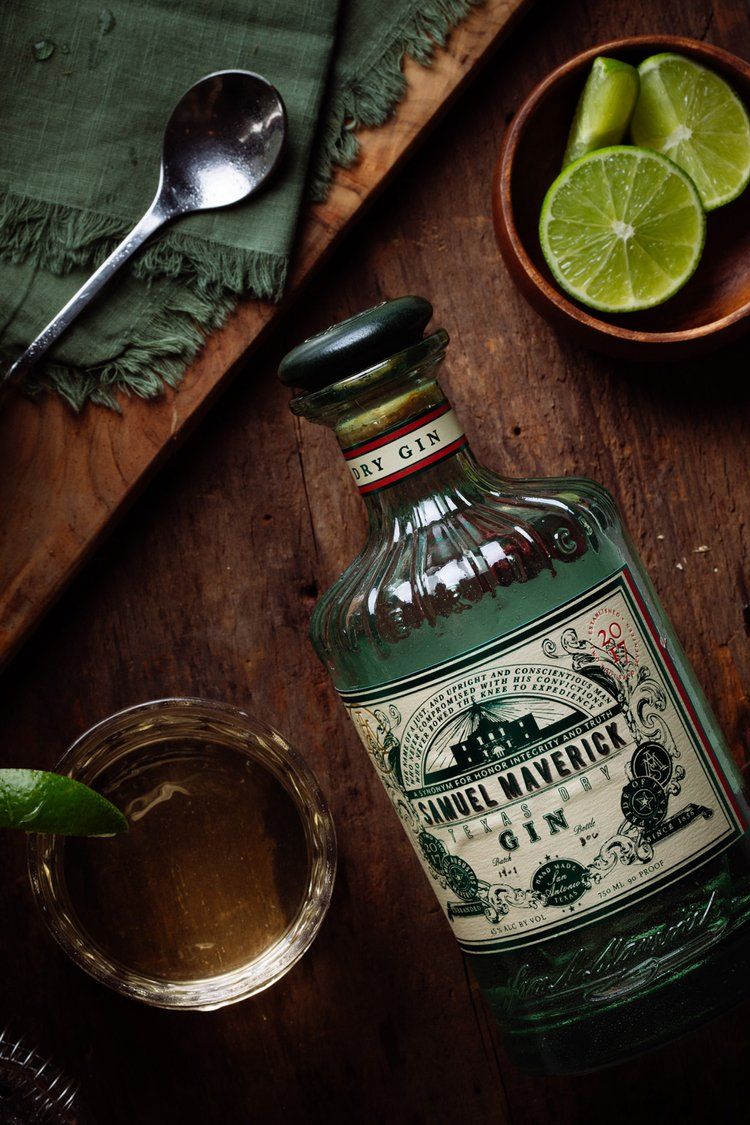 Green glass tequila bottle with vintage label next to sliced limes and cocktail glass on dark wooden surface.