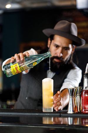 Bartender pouring clear spirits into cocktail glasses at a dimly lit bar counter.