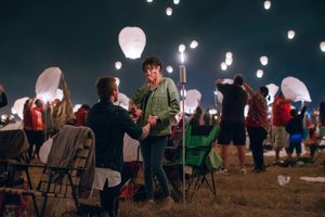 People gather at night to release glowing lanterns into the dark sky while sitting on lawn chairs.