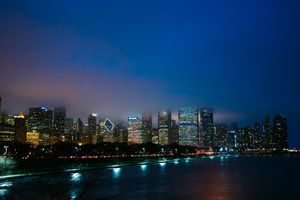 Illuminated Chicago skyline reflects on Lake Michigan at night with glowing city lights against a dark blue sky.