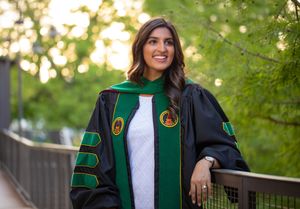 Graduate in academic regalia with green velvet doctoral hood poses in outdoor garden setting.