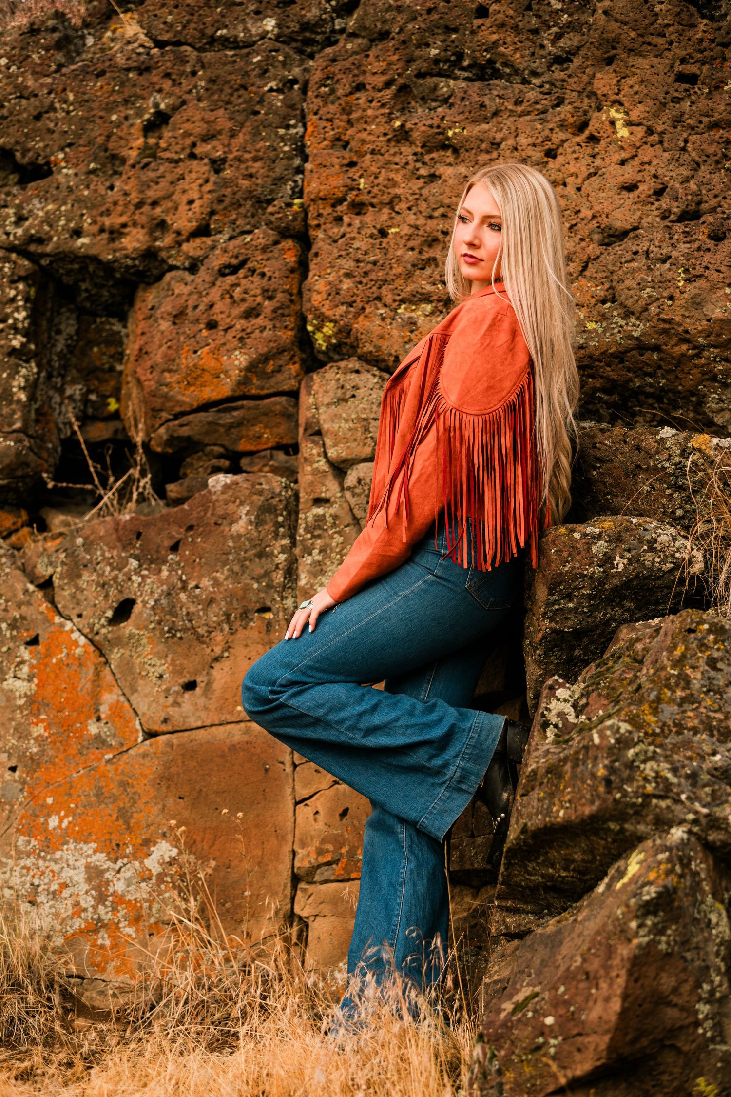 Person in orange fringe jacket and blue denim poses against rustic textured rock wall in outdoor fashion photoshoot.
