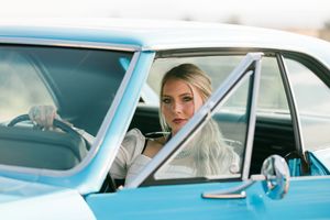 A person sits in the driver's seat of a classic light blue car during a beach-themed photoshoot.