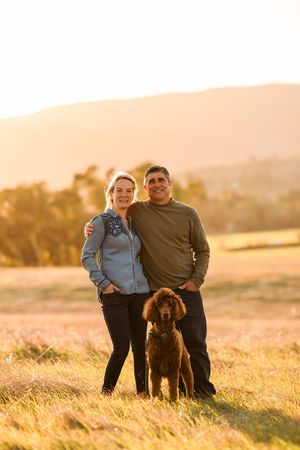 A couple and their brown dog stand together in a golden field at sunset as warm light illuminates the landscape.