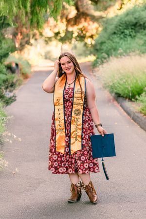 Graduate wearing gold academic stole with burgundy floral dress and cowboy boots poses outdoors during golden hour.