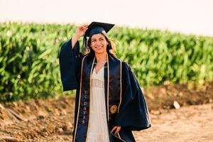 Graduate in blue cap and gown poses outdoors for graduation photos in front of green foliage.