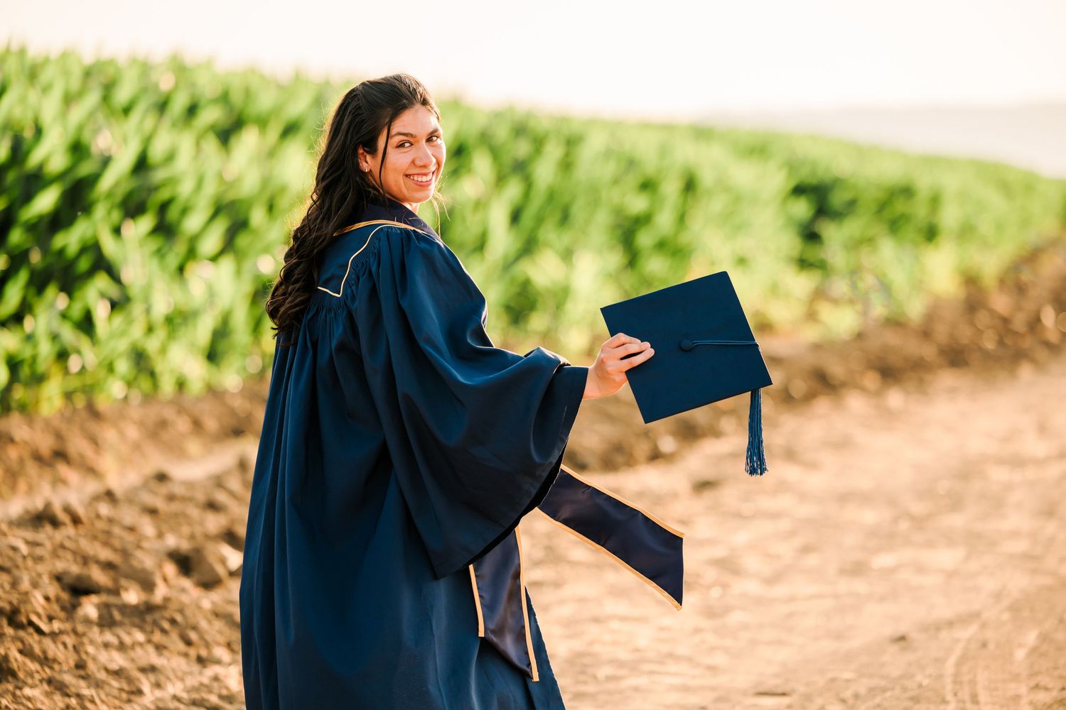 Graduate in navy blue cap and gown walks outdoors while carrying blue diploma cover on sunny day.