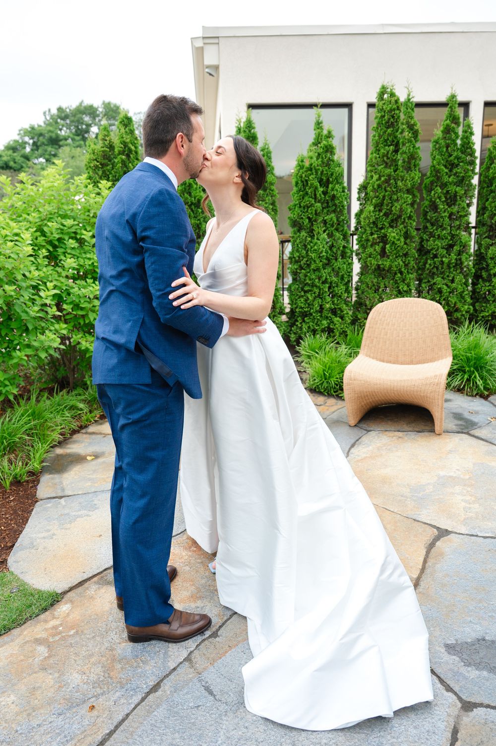 A couple in white and navy blue formal attire share a romantic kiss on a stone patio surrounded by greenery.