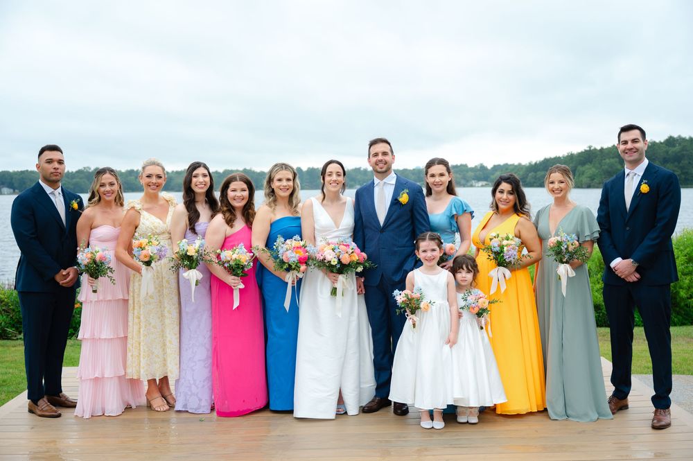 A wedding party in colorful formal attire stands in a row outdoors lakeside at the Lakehouse by Saphire Event Group in Halifax, MA