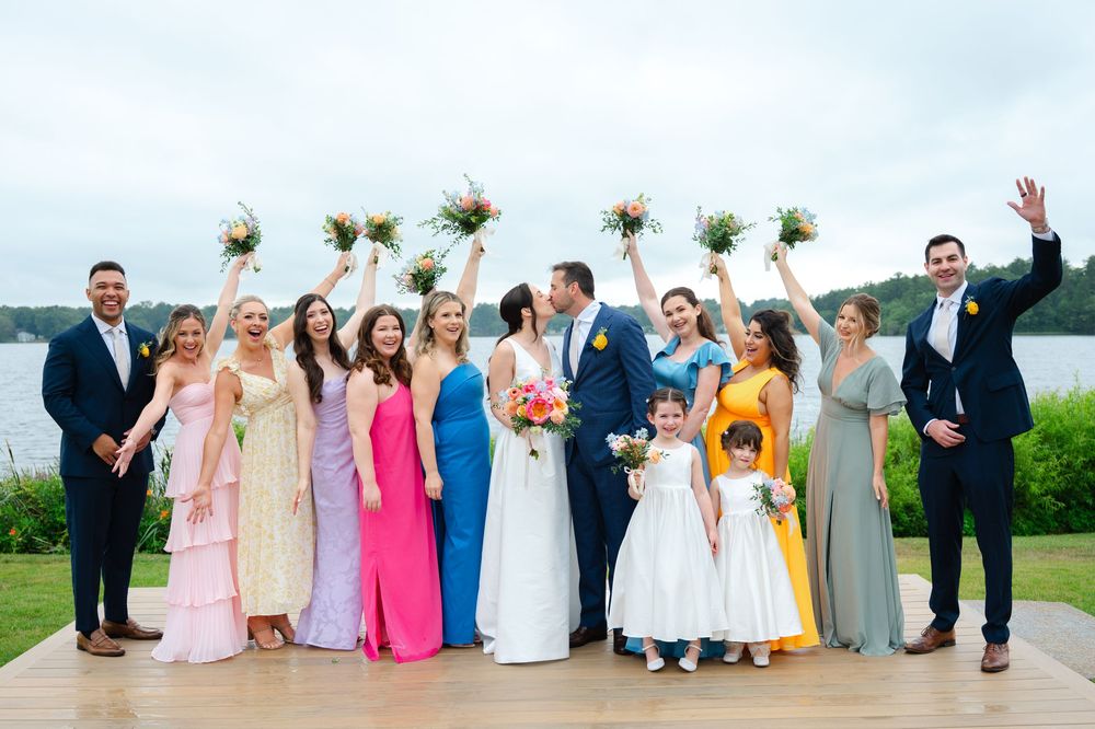 Wedding party in colorful pastel dresses poses together on a grassy hill overlooking the lake at the Lakehouse in Halifax, MA