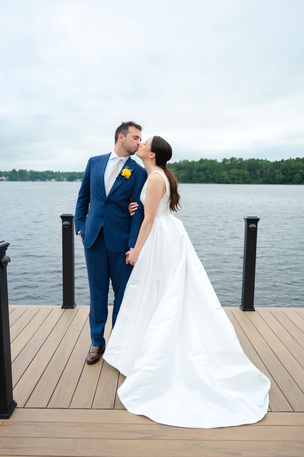 A couple in wedding attire embrace on a wooden dock overlooking a serene lake on their special day at the Lakehouse in Halifax, MA
