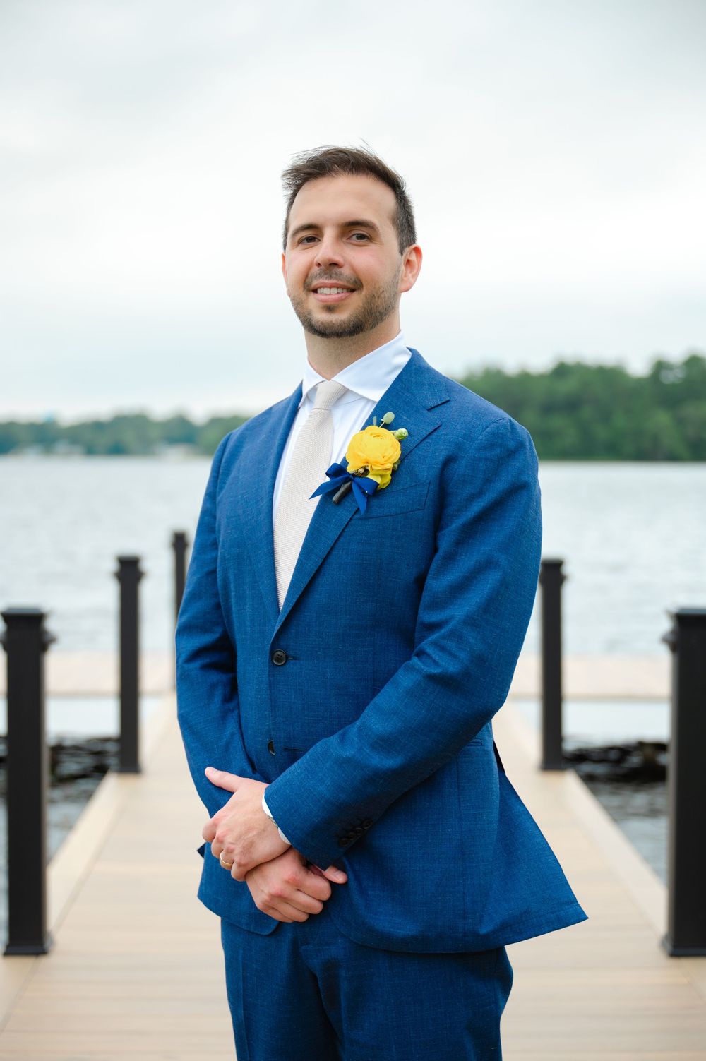 Groom in bright blue suit with yellow boutonniere standing on wooden dock by water.