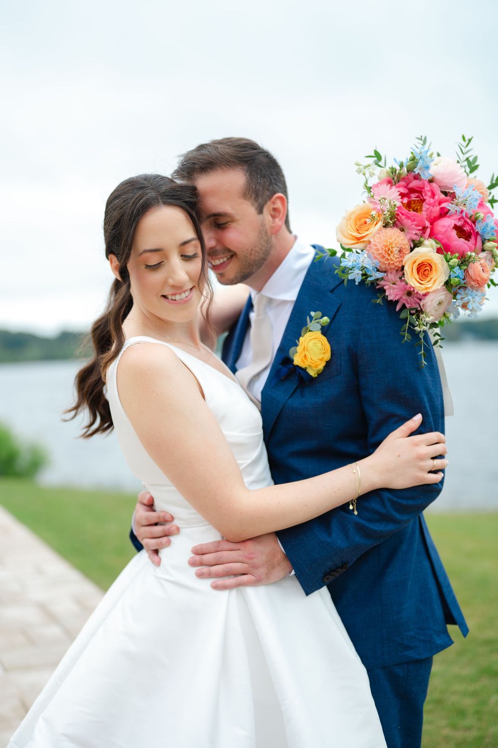 Wedding couple embracing outdoors with vibrant multicolored floral bouquet by the waterfront at the Lakehouse of Saphire Event Group