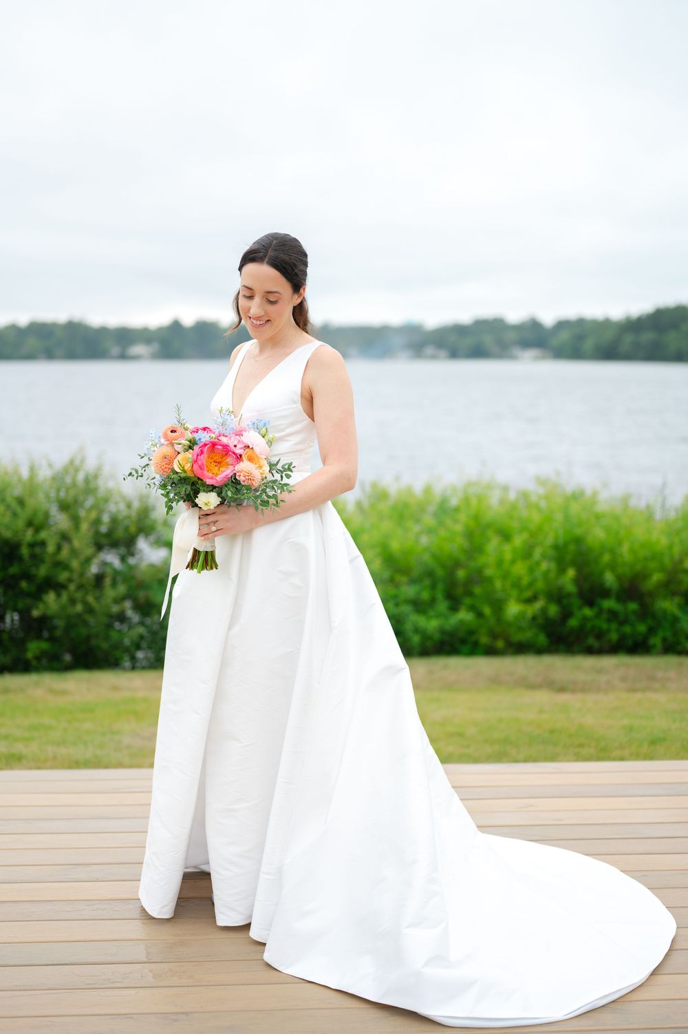 Bride in flowing white wedding gown holding colorful bouquet on wooden deck by waterfront.