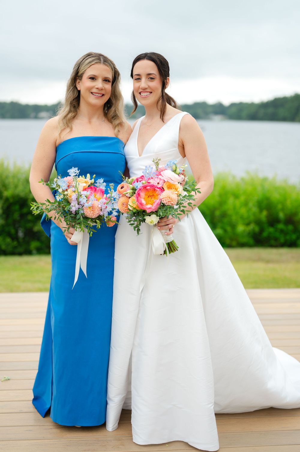 A bride and a bridesmaid pose outdoors in blue and white dresses holding vibrant floral bouquets near a waterfront setting.