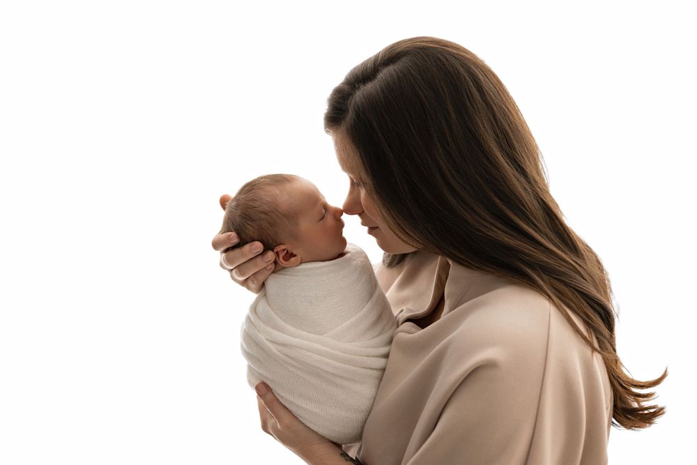 A mother tenderly touches noses with her swaddled newborn baby against a white background.
